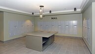 This dedicated mail room is designed to serve a large residential or commercial community with both efficiency and style. The space is lined on three sides with rows of individual, uniform mailboxes finished in a clean light grey, providing ample secure mail storage for numerous occupants. A bold "MAIL" label is prominently displayed on the back wall, offering clear wayfinding.

At the center of the room stands a generously sized sorting island with a smooth, neutral-toned countertop and integrated mail slots, providing a practical surface for organizing and sorting incoming correspondence. The tile flooring in a soft grey tone extends throughout, complementing the overall neutral color palette of the walls and trim.

Overhead, a distinctive cluster pendant light fixture with exposed glass bulbs serves as a subtle design focal point, adding a touch of elegance to the otherwise utilitarian space. Recessed and track lighting supplements the ambient illumination, ensuring the room is well-lit throughout. Crown molding along the ceiling adds a refined architectural detail that elevates the room's overall finish. Together, these elements create a mail room that balances practicality with a polished, well-maintained aesthetic.