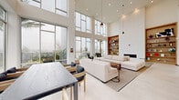Located at 7000 Kiran Patel Dr in Winter Garden, FL, this contemporary two-story amenity space delivers an impressive blend of modern architecture and thoughtfully curated interiors. The main floor welcomes visitors with a dramatic double-height ceiling, floor-to-ceiling glass walls that flood the space with natural light, and light-toned porcelain tile flooring that amplifies the airy atmosphere. A sleek wood-and-glass staircase connects the two levels, anchoring the open layout with refined elegance. The primary living area on this floor features a spacious lounge with plush neutral seating, a built-in fireplace, and a warm wood shelving niche, all framed by sweeping outdoor views of the pool area and surrounding landscape. A charming recessed banquette with gold-toned pedestal tables adds an intimate dining alcove within the otherwise open-plan layout. The upper floor transitions to warm hardwood flooring and offers a more relaxed social environment, complete with comfortable lounge seating arrangements, a full-size billiards table, a ping-pong area, and glass-railed overlooks to the floor below. Large windows on this level provide views of the surrounding community. Recessed lighting throughout both floors ensures a consistent, welcoming ambiance, while the clean white walls and neutral palette lend the entire space a bright, modern character.
