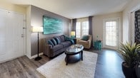 This thoughtfully designed single-floor residence showcases a consistent teal and gray color scheme throughout, creating a cohesive and inviting atmosphere from room to room. The living room serves as the heart of the home, featuring hardwood-style flooring, a bold teal abstract wall painting, and large windows dressed with floor-length drapes that allow ample natural light to fill the space. A plush area rug anchors the seating arrangement, which includes a gray sofa and an accent armchair. The dining area is appointed with a round glass-top table, upholstered chairs, chair-rail molding, and pendant lighting overhead. The kitchen is efficiently laid out with wood-trimmed cabinetry, white appliances including an electric range and refrigerator, and hardwood-style flooring. The primary bedroom features a carpeted floor, a gray accent wall, and generous natural light, while a second bedroom doubles as a flexible workspace with carpet, a sliding glass door, and direct access to a private outdoor patio. The bathroom includes a wood-toned vanity, hardwood-style flooring, and clean, functional finishes. Throughout the residence, decorative artwork, houseplants, and carefully curated accessories contribute to a warm, lived-in character.