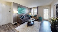 This thoughtfully designed single-floor residence showcases a consistent teal and gray color scheme throughout, creating a cohesive and inviting atmosphere from room to room. The living room serves as the heart of the home, featuring hardwood-style flooring, a bold teal abstract wall painting, and large windows dressed with floor-length drapes that allow ample natural light to fill the space. A plush area rug anchors the seating arrangement, which includes a gray sofa and an accent armchair. The dining area is appointed with a round glass-top table, upholstered chairs, chair-rail molding, and pendant lighting overhead. The kitchen is efficiently laid out with wood-trimmed cabinetry, white appliances including an electric range and refrigerator, and hardwood-style flooring. The primary bedroom features a carpeted floor, a gray accent wall, and generous natural light, while a second bedroom doubles as a flexible workspace with carpet, a sliding glass door, and direct access to a private outdoor patio. The bathroom includes a wood-toned vanity, hardwood-style flooring, and clean, functional finishes. Throughout the residence, decorative artwork, houseplants, and carefully curated accessories contribute to a warm, lived-in character.