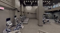 This fitness center presents a comprehensive, multi-zone workout environment designed to accommodate a wide range of training styles. The space is finished in a consistent dark grey palette throughout, with exposed concrete walls, structural columns, and industrial-style exposed piping that contribute to a sleek, urban aesthetic. Overhead string lighting and recessed spotlights provide focused illumination across each zone.

The cardio area is generously sized, housing a full row of treadmills and upright stationary bikes, each equipped with individual display screens. A branded crest logo adorns the wall, reinforcing the facility's identity. An adjacent zone features additional treadmills and recumbent bikes arranged along the perimeter walls.

A dedicated functional training alcove contains an organized rack system stocked with kettlebells, medicine balls in various weights, TRX suspension trainers, and stability balls, offering ample resources for dynamic, bodyweight-based workouts. A separate strength training room houses a cable lat pulldown machine and a flat bench, providing a quieter space for isolated strength work.

The main floor also includes a variety of selectorized and plate-loaded machines arranged across an open layout. A mezzanine level is accessible via a staircase, visible in the main area, and large graphic wall murals of an athletic figure add energy to the overall environment.