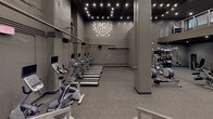 This fitness center presents a comprehensive, multi-zone workout environment designed to accommodate a wide range of training styles. The space is finished in a consistent dark grey palette throughout, with exposed concrete walls, structural columns, and industrial-style exposed piping that contribute to a sleek, urban aesthetic. Overhead string lighting and recessed spotlights provide focused illumination across each zone.

The cardio area is generously sized, housing a full row of treadmills and upright stationary bikes, each equipped with individual display screens. A branded crest logo adorns the wall, reinforcing the facility's identity. An adjacent zone features additional treadmills and recumbent bikes arranged along the perimeter walls.

A dedicated functional training alcove contains an organized rack system stocked with kettlebells, medicine balls in various weights, TRX suspension trainers, and stability balls, offering ample resources for dynamic, bodyweight-based workouts. A separate strength training room houses a cable lat pulldown machine and a flat bench, providing a quieter space for isolated strength work.

The main floor also includes a variety of selectorized and plate-loaded machines arranged across an open layout. A mezzanine level is accessible via a staircase, visible in the main area, and large graphic wall murals of an athletic figure add energy to the overall environment.