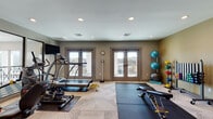 This fitness center is a thoughtfully designed, single-level workout space that accommodates a wide range of exercise needs. The room features a herringbone-patterned carpet flooring that transitions to rubber mat zones, providing dedicated areas for both cardio and strength training activities.

The cardio section is outfitted with a treadmill, elliptical, stair climber, and a recumbent cycling machine, all positioned near a wall-mounted flat-screen television. A large floor mirror on one wall enhances the sense of space while allowing users to monitor their form. Two sets of French doors along one wall provide abundant natural light and open to an outdoor balcony with views of the surrounding landscape.

The strength and functional training area includes a weight bench, multiple resistance machines, a full rack of dumbbells, and a medicine ball storage tower stocked with various sizes. Stability and exercise balls are neatly organized on a vertical rack, and a portable whiteboard stand adds a coaching or programming element to the space.

Accent walls in warm gold and neutral taupe tones add visual depth, complemented by contemporary abstract artwork. Recessed lighting illuminates the entire room evenly. A wall clock and a body measurement station round out the practical amenities, making this a comprehensive and well-appointed fitness facility.