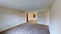 This single-floor residential space offers a straightforward and functional layout with seamlessly connected living areas. The main living space is generously proportioned, featuring neutral wall-to-wall carpeting that creates a warm and cohesive feel throughout. Cream-colored walls paired with light-toned ceilings contribute to a clean, airy ambiance. A subtle step-down or defined transition leads into a secondary living or dining area, which is distinguished by a ceiling fan with integrated lighting, providing both comfort and illumination. A mirrored wall panel in this adjoining space enhances the sense of openness and reflects light effectively, making the area feel more expansive. Wooden interior doors with a rich, dark finish add a classic contrast against the lighter walls and flooring. Tan wood-toned baseboards run along the perimeter, adding a grounding detail to the overall aesthetic. The open sightlines between the connected rooms promote a natural flow and versatile use of the space. Overall, the layout is practical and adaptable, offering a neutral palette that serves as a blank canvas for personalization.