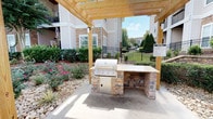 This single-story community space combines sophisticated interior design with thoughtfully crafted outdoor amenities. Inside, the main lounge area makes an immediate impression with soaring ceilings and an abundance of natural light streaming through oversized arched and multi-pane windows that frame views of the surrounding grounds. A classic white-surround fireplace serves as the room's focal point, topped with gold-framed artwork and flanked by light wood-tone flooring that runs throughout. An adjoining sitting area features refined furnishings, including wingback chairs with gold accents, a concrete coffee table, a floor-length gilded mirror, and a lush tropical plant, lending the space an upscale, welcoming atmosphere. The open layout connects seamlessly to a bright, modern kitchen outfitted with stainless steel appliances, white cabinetry, and a decorative tile backsplash. Outdoors, a sturdy wooden pergola shelters a built-in stone outdoor kitchen equipped with a stainless steel gas grill and ample counter space. The surrounding landscape features manicured shrubs, flowering rose bushes, decorative river rock beds, and mature trees, creating a serene and inviting outdoor gathering environment.