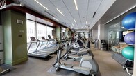 This modern fitness center offers a comprehensive workout environment designed to accommodate a wide range of exercise needs. The cardio zone is prominently featured along one side of the floor, lined with a generous row of treadmills and elliptical trainers from Life Fitness, positioned to take full advantage of the large floor-to-ceiling windows that flood the space with natural light and provide views of the surrounding area. The flooring transitions between rubber matting in high-traffic equipment zones and smooth hard flooring throughout the rest of the space, providing both functionality and comfort. The ceiling is fitted with a clean drop-tile grid accented by linear LED lighting that runs the length of the room, ensuring bright, even illumination throughout. Further into the space, a dedicated strength training and functional fitness area houses a variety of weight machines, free weights, and additional equipment. The layout is thoughtfully organized to allow for easy circulation between stations. Neutral tones in the walls and finishes, combined with dark accent panels, give the facility a polished, professional aesthetic. Stacked folding mats are also visible, suggesting space for stretching or group fitness activities.