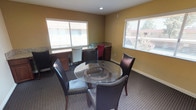 This single-floor space is designed to serve as a versatile meeting and business center, combining collaborative seating with dedicated individual workstations in one cohesive room. The central gathering area features a round glass-top table surrounded by dark leather chairs, providing a comfortable setting for group discussions or informal meetings. The walls are painted in warm neutral tones of cream and tan, contributing to a welcoming and professional ambiance throughout.

Lining the perimeter of the room are rich, dark wood built-in cabinetry units topped with granite countertops, offering ample workspace for individual use. Multiple seating positions at these countertops — including a mix of dark and lighter accent chairs — make the space well-suited for tasks such as computer work or focused individual projects.

Multiple windows fitted with horizontal blinds are distributed around the room, allowing natural light to fill the space while maintaining privacy and light control. Recessed ceiling lighting supplements the natural light, ensuring the room remains well-lit throughout the day. Patterned carpet underfoot adds texture and a grounded feel to the interior. A connecting doorway visible from the main area suggests easy access to adjacent rooms, enhancing the overall functionality and flow of the space.