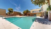 Located at 7834 Viscount Blvd in El Paso, Texas, this outdoor amenity area is centered around a generously sized swimming pool that serves as the focal point of the space. The pool features clearly marked shallow and deeper sections, with no-diving signage, and is framed by a broad concrete sundeck lined with teal lounge chairs — ideal for relaxing under the West Texas sun. Mature trees, including large pines and shade trees, are strategically positioned throughout the grounds, providing natural shade and greenery that softens the open, sun-drenched environment. The surrounding multi-story building features warm yellow stucco exteriors with brick pillar accents and teal-accented doors and railings, lending the space a cohesive, Southwestern aesthetic. An exterior staircase connects levels of the adjacent structure, and a covered walkway area with brick columns provides sheltered access to restroom and utility facilities near the pool. The grounds are enclosed by a teal metal fence with gated entry, ensuring a secure and private atmosphere. Well-maintained landscaping with trimmed shrubs and grassy areas lines the perimeter, contributing to the overall inviting character of this outdoor retreat.