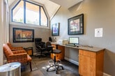 This single-story commercial space in Somerset, NJ, offers a thoughtfully designed environment suited for professional use. The entryway sets a polished first impression with sleek black-framed glass panel doors, wood-look flooring, and a branded welcome mat, leading seamlessly into the interior. Glimpses through the glass reveal multiple functional areas, including what appears to be a fitness room equipped with exercise equipment.

The business center is a standout feature, distinguished by its soaring vaulted ceiling with angular clerestory windows that flood the room with natural light. Built-in wood cabinetry with granite-style countertops lines one wall, accommodating computer workstations and office equipment. Striped upholstered armchairs provide comfortable seating alongside the task-oriented setup.

A private office or consultation area offers a more intimate setting, featuring large windows with outdoor views of lush greenery, colorful framed wall art, and a mix of executive and guest seating. Neutral carpet tile flooring and warm gray walls carry throughout, providing a cohesive, professional aesthetic. Additional service areas with granite countertops and frosted glass doors complete this well-appointed space.