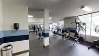 This fitness center offers a spacious, single-floor layout designed to accommodate a wide range of workout needs. The main exercise area features durable rubber flooring throughout, providing both comfort and safety for users. Structural columns are integrated into the space and dressed with a distinctive two-tone blue horizontal stripe detail, complementing the white walls and contributing to a clean, professional aesthetic.

The strength training section is well-appointed with multiple cable and resistance machines, a leg press, and a dedicated free weight area housing a full rack of dumbbells. Large wall-mounted mirrors run along one side of the room, creating a sense of openness while allowing users to monitor their form. The cardio section includes ellipticals, stationary bikes, and treadmills, with a flat-screen television mounted on the wall for entertainment during workouts.

Natural light filters into the space through multiple windows, offering views of the surrounding outdoor greenery. Overhead fluorescent lighting and ceiling fans ensure the environment remains bright and well-ventilated. An on-site restroom provides added convenience, finished with tile flooring, a pedestal sink, and clean, neutral wall tiling. The overall ambiance is functional and well-maintained, making this facility a practical and comprehensive space for fitness activities.