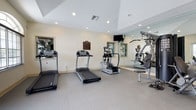 This single-floor fitness room offers a practical and well-organized workout environment designed to support a range of exercise routines. The space is finished with light-colored walls and recessed lighting that creates a bright, welcoming atmosphere conducive to physical activity. The flooring features interlocking tile matting, providing both durability and comfort underfoot during workouts.

The room is equipped with two treadmills positioned side by side, an elliptical trainer for low-impact cardiovascular exercise, and a multi-function strength training machine that accommodates a variety of resistance exercises. A wall-mounted flat-screen television is positioned at a comfortable viewing angle, allowing users to stay entertained during cardio sessions.

Full-length mirrors line one wall, visually expanding the space and allowing users to monitor their form while exercising. A framed informational plaque is mounted on the wall, likely outlining facility rules or guidelines. Natural light filters in through a window, complementing the recessed overhead lighting. The overall layout is clean and uncluttered, making efficient use of the available space while maintaining clear pathways between each piece of equipment.