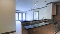 This well-appointed single-floor space showcases a thoughtfully designed open-concept layout that seamlessly connects the kitchen and living areas. The kitchen is a standout feature, boasting rich dark granite countertops that contrast elegantly with warm wood cabinetry finished with contemporary brushed metal hardware. A peninsula-style island provides additional workspace and acts as a natural divider between the kitchen and the adjoining living space. Stylish pendant lights hang above, adding focused illumination and a modern aesthetic touch. The kitchen also features a stainless steel sink and modern fixtures, completing its polished look. A decorative mirror on the kitchen wall enhances the sense of depth and light in the space. The living area benefits from generous natural light streaming through large windows dressed with horizontal blinds, creating a bright and airy atmosphere. A subtle crown molding detail runs along the upper walls, adding a refined architectural element throughout. The neutral color palette, combining clean white walls with warm flooring, lends a versatile and inviting ambiance that is both modern and timeless. Recessed and track lighting throughout ensures well-distributed illumination across the entire space.
