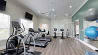 This fitness center offers a clean, well-appointed space designed to support a variety of workout routines. The room features hardwood-style flooring throughout, providing a durable and attractive surface underfoot. Natural light floods the space through multiple windows dressed with white blinds, creating a bright and energizing atmosphere. Two ceiling fans ensure comfortable airflow during workouts, complementing the recessed lighting overhead. Crown molding adds a refined architectural touch to the otherwise functional space.

The cardio area is well-stocked with Matrix-brand treadmills and an elliptical trainer, ideal for sustained aerobic exercise. A row of stationary cycling bikes with integrated display screens occupies the center of the room, offering an engaging cycling experience. A wall-mounted flat-screen television is positioned for easy viewing during workouts. One accent wall is painted in a soft teal tone, adding a subtle pop of color to the otherwise neutral palette of whites and greens. A mirrored partition near the rear of the room enhances the sense of depth and space. The overall ambiance is light, airy, and inviting, making it a welcoming environment for fitness enthusiasts of all levels.
