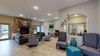 This thoughtfully designed single-floor space exudes a modern, welcoming ambiance through a carefully curated blend of contemporary furnishings and inviting architectural details. The open layout is anchored by a striking floor-to-ceiling stacked stone fireplace with a rich wood mantel, serving as a dramatic focal point and adding warmth to the room. Light wood-toned flooring runs throughout, complementing the neutral grey walls and creating a bright, airy atmosphere enhanced by recessed lighting overhead. The seating arrangement is both functional and stylish, featuring a mix of grey sofas, accent chairs, and teal upholstered ottomans that inject pops of color throughout the space. A decorative arched mirror adorned with sculptural butterfly accents adds an artistic touch near the entryway, while wall art and lush greenery contribute to the overall sense of life and vibrancy. Large windows allow natural light to filter in, further brightening the interior. A secondary seating nook with a distinctive egg-style chair and a sleek console table offers an intimate setting within the larger open space. The overall design strikes a balance between comfort and sophistication, making this a versatile and visually engaging environment suited for relaxation and social interaction alike.