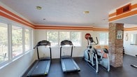 Located at 8424 Madison Ave in Indianapolis, this single-story fitness center offers a well-equipped workout space designed with both function and visual energy in mind. The interior is immediately defined by its bold aesthetic: crisp white walls are accented by two horizontal orange stripes that run continuously around the room, giving the space a dynamic, energetic character. Dark rubber flooring throughout provides a durable, gym-appropriate surface underfoot, while recessed lighting and a textured ceiling ensure consistent illumination across the facility.

The main workout area is generously proportioned, featuring multiple treadmills and an elliptical trainer positioned along a bank of windows that overlook an outdoor pool and landscaped grounds, providing natural light and motivating views during cardio sessions. A structural brick column adds an architectural element to the space, complementing the industrial-style exposed ductwork visible in the secondary area. This adjoining section houses a spin bike, stability balls, a balance board, and a weight bench, along with a dumbbell rack for strength training. Orange seating along the walls provides a rest area between exercises. A wall-mounted mirror enhances the sense of space, and large glass double doors offer direct access to the outdoor amenity area.