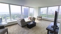 This single-floor living space is defined by its expansive floor-to-ceiling windows that wrap around the room, flooding the interior with abundant natural light and framing dramatic panoramic views of a lush urban park, a shimmering body of water, and a striking city skyline. The generous living area is finished with plush, neutral-toned carpeting that adds warmth and comfort underfoot. The room's light sage-green walls complement the bright, airy atmosphere created by the oversized glazing. A comfortable sectional sofa anchors the seating area, paired with a sleek dark upholstered ottoman that doubles as a coffee table, and an accent chair with a colorful patterned cushion adds a subtle design detail. Perimeter baseboard heating units run beneath the windows, and the open layout allows for flexible furniture arrangement. The overall ambiance is calm and sophisticated, with the sweeping outdoor vistas serving as the space's most compelling architectural feature, making the cityscape and greenery a natural focal point from virtually every angle in the room.