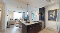 This contemporary high-rise residence is defined by its striking floor-to-ceiling windows, which flood the open-concept living and dining areas with natural light and frame expansive city skyline views. The living room features a corner glass wall with direct balcony access, creating a seamless indoor-outdoor connection. Light hardwood flooring runs throughout the main living spaces and bedrooms, contributing to a warm, cohesive aesthetic.

The kitchen is equipped with dark espresso cabinetry, granite countertops, stainless steel appliances including a built-in microwave and range, and a central island with an undermount sink and pendant lighting overhead. Track lighting supplements the natural light in the main living area.

The primary bedroom offers floor-to-ceiling windows with city views, while a secondary bedroom features softer, neutral tones. Both bathrooms are finished with large-format tile flooring, dark vanity cabinetry, granite countertops, and glass-enclosed showers. The primary bathroom additionally includes a soaking tub and direct access to a walk-in closet. A dedicated laundry room houses a full-size side-by-side washer and dryer with wire shelving above for additional storage.