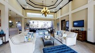 This single-floor clubhouse is a beautifully appointed communal space that blends classic elegance with an open, inviting atmosphere. The focal point of the main lounge is its dramatic multi-tiered tray ceiling, adorned with dark crown molding and recessed lighting, anchored by a statement chandelier that casts a warm, welcoming glow throughout the room. Generous clerestory windows line the upper walls, flooding the interior with abundant natural light and enhancing the sense of grandeur and vertical space. Large retractable glass openings connect the interior lounge seamlessly to an outdoor terrace area, offering unobstructed views of the surrounding community and lush landscaping beyond. The lounge is furnished with plush, neutral-toned sofas accented by blue and patterned throw pillows, centered around a rustic wood coffee table and a tufted navy blue ottoman — a refined yet relaxed seating arrangement suited for both casual gatherings and social events. A dining area is visible toward the rear of the space, providing additional functionality for residents. Dark wood trim details throughout the interior add a touch of traditional sophistication, while the warm beige walls contribute to the overall sense of comfort and refinement that defines this clubhouse.