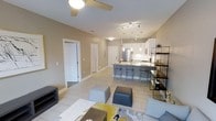 This single-floor residence showcases an open-concept layout that integrates the living, dining, and kitchen areas into one cohesive space. The kitchen is a standout feature, equipped with white cabinetry, stainless steel appliances, and a central island with bar seating, illuminated by elegant pendant lights that hang at varying heights. Track lighting above the kitchen adds focused illumination, complementing the warm overhead ceiling fan light in the living area.

The living space is finished with light-toned tile flooring throughout, contributing to a clean, airy aesthetic. Neutral warm beige walls provide a versatile backdrop, while the contemporary furnishings — including modular ottomans and a patterned area rug with yellow and gray accents — add personality and color. A sleek open shelving unit along one wall offers both display and storage functionality.

Doorways visible from the main living area suggest additional rooms, maintaining a sense of privacy while keeping the central space open and social. The overall ambiance is modern and well-appointed, balancing practicality with a welcoming, stylish atmosphere.
