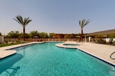 Located at 870 S Sarival Ave in Goodyear, Arizona, this single-level outdoor community amenity area is designed to deliver a resort-style experience in the heart of the Sonoran Desert. The centerpiece is a generously sized freeform swimming pool with crystal-clear turquoise water, complemented by an attached circular spa that provides a relaxing retreat. The expansive pool deck is finished in light-toned paver stone, creating a clean and inviting surface that surrounds the water on all sides. Towering date palms are strategically placed throughout the space, offering natural shade and a distinctly desert aesthetic. Multiple seating zones are distributed around the deck, including lounge chairs positioned along a stretch of artificial turf — a cool, comfortable alternative to hard paving. Umbrella-shaded tables and chairs provide additional gathering areas for socializing or dining. A covered pergola structure anchors one end of the space, offering a shaded retreat for residents seeking relief from the Arizona sun. An outdoor kitchen or grill station is also present, enhancing the space's suitability for community gatherings. The perimeter is enclosed by iron fencing with lush greenery beyond, providing privacy while maintaining an open-air atmosphere. Accessibility features, including a pool lift, are visible, reflecting an inclusive design approach throughout this well-appointed community outdoor space.