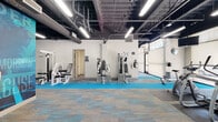 This fitness center is designed to support a comprehensive workout experience across a single, open-plan floor. The space features an industrial-style aesthetic, with an exposed ceiling showcasing dark-painted ductwork, structural elements, and suspended linear LED lighting that keeps the entire facility brightly and evenly illuminated.

The main workout floor is finished with a distinctive multi-tone carpet tile in blue, gray, and tan, accented by vibrant blue runner strips that define exercise zones throughout the space. Large floor-to-ceiling windows along one perimeter wall allow ample natural light to filter in, offering views to the exterior and contributing to an energetic, open atmosphere.

Cardio equipment includes treadmills, ellipticals, stationary bikes, and a rowing machine, positioned to take advantage of both the natural light and wall-mounted televisions. A dedicated strength training zone houses multiple selectorized resistance machines, while a functional training area features a Life Fitness Synrgy360 multi-station rig equipped with battle ropes, along with stability balls, kettlebells, and a dumbbell rack.

Bold motivational wall murals in deep blue tones serve as striking focal points throughout the space, reinforcing an energetic and goal-oriented atmosphere. A separate stretching and accessory area provides space for warm-ups, cool-downs, and group exercise. Full-length mirrors along key walls enhance the sense of depth and allow users to monitor their form during workouts.