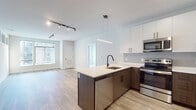 This well-appointed single-floor unit offers a thoughtfully designed open-concept layout that seamlessly connects the kitchen and living area. The kitchen is a standout feature, showcasing two-tone cabinetry with rich dark lower cabinets and crisp white upper cabinets extending to the ceiling for ample storage. A large peninsula with a light quartz countertop and undermount sink with a matte black faucet serves as both a functional workspace and a social hub. Stainless steel appliances, including a refrigerator, electric range, and built-in microwave, complete the kitchen. Light wood-look flooring flows throughout the main living areas, complementing the neutral palette and contributing to a bright, airy atmosphere. Track lighting and modern pendant fixtures provide layered illumination. The living area is generously proportioned with clean white walls and large windows that invite natural light. The bathroom features a floating vanity with wood-tone cabinetry, black hardware accents, a framed mirror, and a glass-enclosed shower. The bedroom is finished with plush carpet and connects to a walk-in closet outfitted with wire shelving. Black hardware accents on doors and fixtures provide a cohesive, contemporary touch throughout the space.