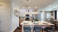 This thoughtfully designed single-floor residence blends contemporary style with functional living. The open-concept kitchen is a standout feature, showcasing crisp white upper cabinetry paired with warm wood-toned lower cabinets, a large center island with quartz countertops and bar seating, stainless steel appliances, and sleek pendant lighting that adds a modern touch. Rich dark hardwood flooring flows throughout the main living areas, anchoring the space with warmth and sophistication.

The living room is bright and inviting, featuring multiple windows dressed with layered blinds and dark curtain panels that allow for flexible light control. A ceiling fan and curated artwork contribute to the room's polished ambiance.

The primary bedroom is generously sized with plush carpet, a tray ceiling, and natural light streaming through large windows. It connects to a well-appointed en-suite bathroom featuring a dual-sink vanity with wood cabinetry, quartz countertops, and a walk-in closet. A second bedroom offers similar comfort with soft carpet and a soothing accent wall.

An additional bathroom echoes the same refined aesthetic, with a wide vanity, large mirror, and warm wood finishes. Throughout the residence, consistent design elements—neutral tones, wood accents, and contemporary fixtures—create a cohesive and welcoming atmosphere.