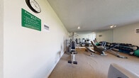 The fitness center at 13222 E Iliff Ave in Aurora, Colorado, is a dedicated exercise facility housed within a single, efficiently organized room. The space is outfitted with a comprehensive selection of equipment designed to support a full range of workout routines. Cardio options include multiple treadmills and elliptical trainers, providing ample availability during peak hours. Strength training is well-represented with a Vectra multi-station cable machine, a weight bench, and a dumbbell rack stocked with a variety of free weights. Stability balls are also available for supplementary fitness work. The room features neutral-toned carpet flooring and textured ceilings, contributing to a clean, functional atmosphere. Large wall-to-wall mirrors along one side of the room visually expand the space and allow users to monitor their form during workouts. A floor-to-ceiling window with vertical blinds invites natural light and offers an outdoor view, adding a bright, open quality to the environment. Wall-mounted track lighting provides additional illumination throughout. A wall clock and posted exercise room rules keep the space practical and user-friendly. The overall design prioritizes functionality, making it a convenient and accessible fitness resource.