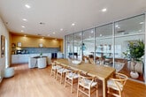 Located at 1512 Hollowhill Drive in Bryan, Texas, this single-floor space offers a thoughtfully designed open-concept dining room and kitchen area. The dining area is anchored by a large natural wood communal table surrounded by classic wishbone chairs, ideal for gatherings or collaborative settings. A wall-mounted display screen adds a modern, functional touch, while floor-to-ceiling glass partition walls flood the space with natural light and provide clear sightlines to adjacent areas. Warm-toned hardwood flooring runs throughout, lending a cohesive and inviting aesthetic. Recessed lighting further brightens the interior. The kitchenette is sleek and efficient, featuring continuous white lower cabinetry topped with smooth countertops, a textured grey tile backsplash, and natural wood upper cabinetry with integrated accent lighting. Two stainless steel beverage coolers are built into the cabinetry, offering practical refreshment storage. A small seating area with a round table and curved chairs adds a casual breakout zone. Together, the clean lines, neutral palette, and mix of warm wood and cool grey tones create a contemporary, professional ambiance well-suited for both social and functional use.