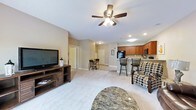 This single-floor residence offers an open-concept layout that seamlessly connects the living, dining, and kitchen areas into one cohesive and functional space. The living room is anchored by plush carpeting, a ceiling fan with integrated lighting, and recessed ceiling lights that provide ample illumination. A media console with a large flat-screen television serves as a focal point, while a marble-topped coffee table and patterned accent chair add character and comfort to the space. Adjacent to the living area, a compact dining nook features a small table and chairs, ideal for casual meals. The kitchen is well-equipped with warm-toned wood cabinetry, black appliances including a refrigerator and microwave, and a raised breakfast bar with bar stool seating — perfect for additional dining or entertaining. Neutral-toned walls and light carpeting throughout create a clean, cohesive aesthetic that enhances the sense of openness. Doorways visible from the main area suggest additional private rooms, contributing to a practical and complete floor plan. The overall ambiance is warm, inviting, and efficiently designed for everyday living.