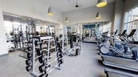 This modern fitness center offers a thoughtfully designed workout environment outfitted with a wide variety of exercise equipment to accommodate diverse training needs. The strength training area is prominently featured, housing multiple barbell racks loaded with weight plates, a cable and Smith machine setup, and organized free weight stations — providing everything needed for a comprehensive resistance training session. Cardio equipment, including elliptical machines, lines the perimeter to complement the strength-focused layout. Large mirrors span the walls, amplifying the sense of space and allowing users to monitor their form throughout their workout. The interior is finished with a neutral-toned carpet floor that softens the space, while the walls are accented with a crisp combination of white and cool blue tones that contribute to a clean, energizing atmosphere. Overhead, stylish pendant lights with warm yellow interiors hang at varying heights, providing ample and inviting illumination throughout. The elevated ceiling height adds an open, airy feel to the space. Overall, the facility strikes a balance between functional design and modern aesthetics, making it a well-appointed destination for fitness enthusiasts of all levels.
