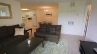 This single-floor space is designed around an open-concept floor plan that seamlessly connects the living area, dining space, and kitchen. The living room serves as the central hub, featuring plush carpeting, a patterned area rug, and a dark-upholstered sofa accented with decorative pillows, creating a welcoming and relaxed atmosphere. A dark-finished coffee table anchors the seating area, complementing the overall color scheme.

Flowing naturally from the living room, the dining area is illuminated by a elegant chandelier that adds a touch of refinement to the space. A modest dining table with place settings is positioned beneath the fixture, making it an inviting spot for meals.

The kitchen is visible from both the living and dining areas, featuring white cabinetry, white appliances including a full-sized refrigerator, and light countertops. The open sightlines between all three areas enhance the sense of spaciousness throughout the unit.

Wall-mounted HVAC vents are integrated into the neutral-toned walls, ensuring climate control throughout the space. The overall ambiance is warm, functional, and practical, with a cohesive neutral palette tying each area together.