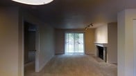 This single-floor space presents a living area characterized by a neutral, understated aesthetic. The room is finished with wall-to-wall carpeting in a warm beige tone, complemented by smooth, neutral-colored walls that create a cohesive and calm atmosphere. A notable architectural detail is the dark-painted ceiling, which contrasts with the lighter walls and adds a distinctive visual element to the space.

A brick fireplace with a white mantel is positioned along one wall, serving as a natural focal point in the room. It features a dark firebox surround, lending the space a traditional yet grounded character. On the opposite side of the room, a sliding glass door fitted with vertical blinds allows natural light to filter into the space and provides a view of surrounding greenery outdoors, offering a connection to the exterior environment.

Track lighting is mounted near the fireplace wall, providing adjustable, directed illumination to supplement the natural light. An additional circular ceiling fixture is visible, contributing to overall lighting. A doorway on the left side of the room leads to an adjacent area, suggesting further rooms within the layout of this single-level space.