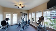 The fitness center at 1845 N Broadway in Escondido is a well-appointed, single-floor exercise space designed to support a variety of workout routines. The room features neutral-toned walls and dark carpeted flooring, creating a clean and functional environment. A ceiling fan with a built-in light fixture keeps the space comfortable during workouts.

Natural light is a standout feature of this fitness center, with multiple large, latticed windows lining the walls and offering views of the surrounding outdoor greenery. A glass-paned door provides additional light and convenient access to the exterior.

The space is equipped with a multi-station strength training machine, ideal for a full range of resistance exercises, as well as cardio equipment including an elliptical trainer. The layout allows for comfortable movement between machines without feeling cramped. The combination of ample natural light, ventilation from the ceiling fan, and a practical equipment selection makes this fitness center a functional and inviting space for residents looking to maintain an active lifestyle.