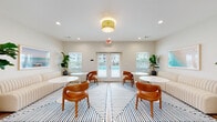 Located at 1200 Deana Rd in Ocean Springs, Mississippi, this single-floor clubhouse facility offers a thoughtfully designed communal space built for both relaxation and activity. The entry foyer welcomes visitors with light wood-look flooring, crisp white walls, and a warm rattan flush-mount light fixture, setting an airy, coastal tone throughout. French doors at the entrance and within the lounge flood the interior with natural light and provide direct views of the outdoor pool area.

The central lounge is spacious and elegantly appointed, featuring creamy ribbed banquette seating, cognac leather accent chairs, and round white tables arranged atop a bold navy-and-white geometric area rug. Lush fiddle-leaf fig and tropical potted plants add life and color, while coastal-inspired artwork adorns the walls. Recessed lighting complements the statement ceiling fixture, ensuring the space is well-lit throughout the day.

A dedicated fitness room is equipped with cardio machines, a multi-function cable weight system, and rubber flooring, offering residents a convenient on-site workout space. A coffee and refreshment station is conveniently located near the entry corridor. The layout is efficient and open, with multiple doorways connecting key areas of the facility seamlessly.