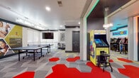 Located at 10051 Perimeter Pkwy in Charlotte, NC, this single-floor amenity space is designed as an energetic hub for entertainment and socializing. The interior is defined by a bold, contemporary aesthetic, anchored by eye-catching hexagonal patterned carpeting in shades of yellow-green, red, and gray that flows throughout the entire space.

The gaming area is a standout feature, housing classic arcade cabinets including a Pac-Man machine and additional retro-style units, each framed by colorful LED accent lighting in green and orange hues. A dedicated VR gaming station adds a modern dimension to the entertainment offerings. A full-size ping-pong table anchors a separate activity zone, accompanied by a wall-mounted flat-screen television ideal for group viewing.

A comfortable lounge area transitions seamlessly from the game zones, featuring modular gray sofas with accent pillows, a wood-topped coffee table, and a sculptural pendant light fixture. Large windows allow natural light to filter in, offering views of the surrounding landscape.

A compact kitchenette supports the space with light wood cabinetry, a stainless steel farmhouse sink, full-size refrigerator, microwave, and an additional mounted television. Exposed concrete columns and dark accent trim add an industrial edge, while recessed lighting throughout ensures a well-lit, welcoming atmosphere.