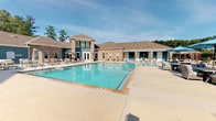 Located at 82 Ardmore Gardens Dr in Hardeeville, South Carolina, this single-story community amenity center serves as a social and recreational hub for the Ardmore Gardens residential community. The centerpiece of the facility is a generously sized resort-style swimming pool with a spacious concrete surround, accommodating lounge chairs for sunbathing and relaxation under the open South Carolina sky. The pool area is framed by a classic brick and blue-accented clubhouse building featuring large windows, French doors, and a pitched roof, lending a polished, welcoming aesthetic. Access to the pool deck is controlled through a black wrought iron security gate set within brick columns, complemented by manicured landscaping including ornamental grasses and palm trees that reinforce the coastal character of the region. A covered outdoor patio extends from the clubhouse, furnished with wicker seating, dining sets, and decorative planters, providing a shaded gathering space with open views of the surrounding community and tree line. An additional outdoor lounge area offers comfortable striped cushioned furniture ideal for casual relaxation. The overall ambiance is bright, airy, and inviting, combining durable brick construction with vibrant coastal color accents to create an attractive and functional outdoor living environment for residents.