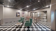 Located at 349–399 Trumbull St in Hartford, CT, this single-floor amenity space combines a retro-inspired lounge area with a fully equipped laundry facility. The space is unified by a striking black-and-white checkered tile floor that runs throughout, creating a bold, cohesive aesthetic. A standout architectural feature is the textured geometric ceiling finished in matte black, lending the space a sophisticated, design-forward character with recessed lighting adding warmth.

The lounge area is furnished with tufted green armchairs arranged around a round marble-top coffee table, complemented by high-top bar seating with blush-toned chairs. Vintage-style French advertisement posters decorate the walls, reinforcing the retro ambiance. A glass-partitioned enclosure with black steel framing separates a dedicated bar-height seating area while maintaining visual openness. Vending machines and a service counter are conveniently positioned near the entrance.

The laundry room continues the same design language with matching checkered flooring and geometric ceiling tiles. It is well-equipped with multiple front-loading washers and dryers, stacked units, and dedicated detergent dispensers, providing ample capacity. Together, the two areas form a functional yet visually engaging communal space.