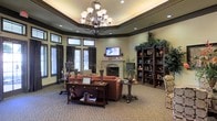 This sophisticated single-floor space exudes warmth and elegance through its thoughtfully curated design. The centerpiece of the room is a striking tray ceiling adorned with dark crown molding, which frames a grand wrought-iron chandelier with cream glass shades. Recessed lighting throughout enhances the ambient glow, complementing the inviting atmosphere.

The living area is anchored by a classic fireplace with a decorative mantel, above which a wall-mounted flat-screen television is prominently displayed. Rich terracotta-toned sofas and patterned armchairs are arranged around a dark wood console table and round side tables, creating a comfortable and cohesive seating arrangement.

A series of tall windows dressed in dark curtains with transom windows above allow natural light to filter into the space, while maintaining a sense of privacy. Lush greenery and decorative plants are positioned throughout, adding a natural element to the refined interior.

Built-in and freestanding dark wood bookshelves line one wall, filled with decorative objects, books, and plants that add depth and visual interest. The carpeted flooring in a neutral tone ties the space together, contributing to the overall sense of comfort and sophistication. This space is well-suited for relaxation, socializing, or quiet reading.