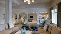 This single-story space is designed with an open, flowing layout that connects multiple functional areas through a series of graceful arched openings, lending the interior a classic and refined architectural character. The main lounge area is generously proportioned, furnished with neutral-toned wingback chairs, upholstered sofas, and tufted ottomans arranged atop patterned area rugs, creating an inviting and comfortable gathering environment. Recessed lighting and a statement multi-pendant chandelier illuminate the space warmly, complementing the soft, neutral wall tones throughout. Beyond the arched passages, a dedicated game room is visible, centered around a billiard table beneath a wall-mounted flat-screen television, offering a casual recreational retreat. To one side, a well-equipped kitchen features white cabinetry, stainless steel appliances, pendant lighting, and a raised counter with bar-height seating—ideal for casual dining or entertaining. The cohesive design palette of creams, taupes, and soft teal accents ties the entire space together, projecting a polished yet welcoming ambiance suited for socializing and relaxation.