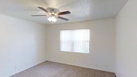 This single-floor space features a well-appointed room finished with wall-to-wall carpeting that adds warmth and comfort underfoot. A central ceiling fan with an integrated light fixture provides both air circulation and ambient lighting, making the room practical year-round. A generously sized window dressed with vertical blinds allows natural light to filter in while offering privacy and light control. The walls are painted in a neutral tone, complementing the textured popcorn ceiling and creating a clean, understated aesthetic throughout. The room's layout is open and versatile, accommodating a variety of furniture arrangements to suit different needs. White baseboards provide a crisp finishing detail along the perimeter of the room. The overall ambiance is bright, quiet, and functional, with a simple design that offers a blank canvas for personalization. The combination of soft carpeting, ample natural light, and ceiling fan creates a comfortable and livable environment suited for everyday use.