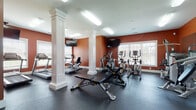 This fitness center at 15201 Flats Dr. in Noblesville, Indiana offers a comprehensive and well-equipped workout facility designed to support a wide range of exercise routines. The space is immediately distinguished by its bold terracotta-orange accent walls paired with crisp white trim and decorative white columns, creating an energetic yet polished atmosphere. Durable rubber flooring spans the entire space, providing a safe and functional surface for all types of training.

The cardio zone features Matrix treadmills, elliptical trainers, and a recumbent bike, all positioned near windows fitted with white blinds that allow natural light to filter in while offering views of the surrounding landscape. Wall-mounted flat-screen televisions are positioned throughout the space for entertainment during workouts.

A dedicated strength-training area includes a multi-station cable machine and a variety of weight benches, while a large wall-spanning mirror enhances the sense of openness and allows users to monitor their form. The free weights section offers a full dumbbell rack alongside barbells, exercise balls in multiple sizes, resistance accessories, and yoga mats neatly organized on a storage rack. Bright overhead fluorescent lighting ensures the entire space is evenly illuminated. The single-floor layout keeps all amenities conveniently accessible in one open, functional environment.