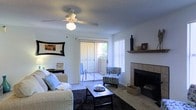 This single-floor residence offers a well-organized layout with clearly defined living spaces visible throughout. The living room serves as a central gathering area, featuring a tile-surround fireplace with a wooden mantel shelf, carpeted flooring, and a ceiling fan with integrated lighting. A sliding glass door opens to a private balcony, allowing natural light to fill the room through vertical blinds, creating a bright and airy atmosphere. The space comfortably accommodates a seating arrangement centered around a coffee table, with room for accent chairs near the fireplace.

Adjacent to the living room, the layout includes a dedicated dining area furnished with a table and chairs, suitable for everyday meals or entertaining. The kitchen is positioned nearby, offering a functional workspace with cabinetry and appliances. A bedroom is also present within the floor plan, providing a private retreat separate from the main living spaces.

The overall layout flows cohesively from one room to the next, with consistent neutral-toned walls and carpeted flooring tying the spaces together. The combination of natural light, a fireplace, and balcony access contributes to a comfortable and inviting ambiance throughout the residence.