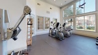 Located at 3620 Huffines Blvd in Carrollton, Texas, this well-appointed fitness center offers a bright, spacious environment designed to support a full range of workout routines. The facility is characterized by soaring ceilings and oversized windows that flood the space with natural light while providing pleasant views of the surrounding landscaped grounds. Ceiling fans are mounted throughout, ensuring comfortable airflow during workouts.

The cardio zone is equipped with multiple treadmills and elliptical trainers, positioned directly in front of large windows and wall-mounted televisions, creating an engaging exercise experience. The strength training area features a comprehensive dumbbell rack, adjustable weight benches, cable machines, and multi-functional strength equipment, all set against a floor-to-ceiling mirror wall that enhances the sense of openness and allows for form monitoring. Additional equipment includes rowing machines and cable/pulley systems.

Durable rubber flooring covers the workout areas, offering both safety and comfort. French-style glass doors provide access to the exterior, further connecting the interior to the outdoor environment. The clean, white-painted walls and recessed lighting contribute to a fresh, modern aesthetic. The layout is thoughtfully organized to separate cardio and strength zones, allowing users to move efficiently through their routines in a well-maintained, welcoming facility.