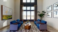 This thoughtfully designed single-floor space functions as a polished communal hub, combining multiple distinct zones within an open, airy layout. The lounge area immediately draws attention with its dramatic floor-to-ceiling windows framed by flowing linen drapes, flooding the space with natural light and offering views of the surrounding landscape. Beneath the towering ceilings, blue upholstered armchairs are arranged symmetrically around an organically shaped copper-toned coffee table atop a layered area rug, creating an inviting conversational setting. A gallery wall featuring a curated grid of six bold abstract artworks anchors a secondary seating nook, adding a strong visual focal point alongside a large tropical potted plant.

The dining area transitions seamlessly from the lounge, offering both a long communal table with rust-colored upholstered chairs and high-top bar tables positioned beside large windows — ideal for casual collaboration or focused work. A large-scale painting brings additional artistic character to this zone.

A dedicated beverage station rounds out the space, featuring dark cabinetry with granite countertops, a coffee machine, sink, and a textured tile accent wall with a wall-mounted television — delivering both function and visual interest. Light stone tile flooring unifies the entire space.