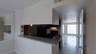This single-floor home offers a functional and cohesive layout centered around an open-concept design that seamlessly connects the kitchen and living areas. The kitchen is a standout feature, equipped with black appliances including a range and refrigerator, complemented by granite countertops that extend along a peninsula-style breakfast bar. A dark mirrored backsplash adds a sleek, modern contrast to the white cabinetry, while under-cabinet lighting provides a warm, practical glow. A HVAC vent positioned above the kitchen ensures consistent climate control throughout the space. The kitchen flows naturally into the adjoining living area, which benefits from a bright window that draws in natural light, creating an open and airy atmosphere. Light-toned tile flooring runs throughout, offering a clean and uniform look that ties the spaces together. The textured ceilings add a classic architectural detail, while white walls keep the interior feeling bright and neutral. A fire extinguisher is conveniently mounted near the kitchen, reflecting thoughtful safety considerations. Overall, this home presents a straightforward, well-appointed layout suited for comfortable everyday living.
