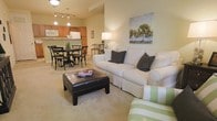 This single-floor space showcases an open-concept floor plan that seamlessly connects the living, dining, and kitchen areas into one cohesive and functional environment. The living area features plush wall-to-wall carpeting in a warm neutral tone, providing a comfortable foundation for the space. A white sofa accented with dark throw pillows anchors the seating arrangement, complemented by a dark leather ottoman that doubles as a coffee table and a green-striped accent chair, adding a touch of color. Decorative wall art featuring a nature-inspired tree motif adds visual interest above the sofa.

The dining area transitions naturally from the living space and accommodates a round dark-wood table surrounded by classic cross-back chairs, illuminated overhead by a stylish pendant light fixture. Beyond the dining area, the kitchen features warm wood cabinetry, light countertops, and standard appliances including a refrigerator and microwave, with track lighting providing functional illumination. The open layout and consistent neutral color palette throughout create a cohesive, inviting ambiance, making this space ideal for both everyday living and casual entertaining.