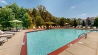 Located in the Irene Woods community of Collierville, Tennessee, this single-story amenity space offers residents a well-designed outdoor retreat with multiple gathering areas. The centerpiece is a generously sized swimming pool with a curved edge and brick coping detail, its clear blue water framed by mature trees that provide a lush, natural backdrop and a sense of privacy.

The expansive concrete sundeck surrounding the pool is furnished with rows of adjustable lounge chairs shaded by bold yellow-green cantilever umbrellas, creating an inviting environment for relaxation. A separate outdoor lounge area features a rustic wooden pergola sheltering cushioned seating, offering a more intimate gathering spot alongside manicured landscaping and ornamental trees.

The adjacent clubhouse building is architecturally distinguished, combining classic brick and white-painted siding with a steeply pitched roofline, dormer windows, and large multi-pane windows that flood the interior with natural light. The building's arched brick entry and covered porch add to its traditional character. The entire outdoor area is enclosed by a sleek black iron fence, providing security while maintaining open, welcoming sightlines throughout the community.