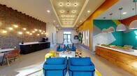 Located at 1101 S Beechwood Ave in Fayetteville, Arkansas, this dynamic single-floor communal space makes a bold first impression with its energetic design and thoughtfully curated interiors. The open-plan layout is anchored by a striking amber-yellow accent wall adorned with geometric moss art installations and illuminated marquee lettering, creating an eye-catching focal point upon entry. A vivid mural with teal and orange geometric patterns adds additional artistic depth to the space.

The reception area features a clean white counter with wood-paneled detailing, complemented by a multicolored mosaic tile floor that adds playful character underfoot. A central lounge zone is furnished with bold cobalt blue armchairs, yellow ottoman seats, and a round white coffee table, all arranged atop a graphic patterned area rug.

A full-height exposed brick wall runs along one side of the space, softened by cascading globe pendant lights that cast a warm, ambient glow. Adjacent to the brick wall, a casual seating nook offers plush light blue modular sofas with accent pillows and marble-topped side tables — an inviting spot for informal gatherings. Large windows throughout flood the interior with natural light and provide views of the surrounding Fayetteville streetscape. A coffee and refreshment station with dark cabinetry completes the thoughtfully appointed amenity space.