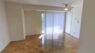This single-floor space is highlighted by its distinctive parquet wood flooring, which adds warmth and visual interest throughout the main living area. The floors are polished to a reflective sheen, contributing to a bright and clean aesthetic. A ceiling fan with an integrated light fixture is centrally positioned overhead, providing both air circulation and ambient lighting.

The room is anchored by a full-width sliding glass door dressed with vertical blinds, allowing for adjustable natural light and direct access to an outdoor area. Glimpses of greenery are visible through the door, suggesting a pleasant outdoor setting. The neutral, light-toned walls complement the warm flooring and keep the space feeling open and airy.

A door on the adjacent wall provides passage to another area of the unit. The structural detailing includes a visible ceiling beam element that adds subtle architectural definition. Overall, the space has a functional, well-maintained character with a balance of natural materials and practical features suited for everyday living.